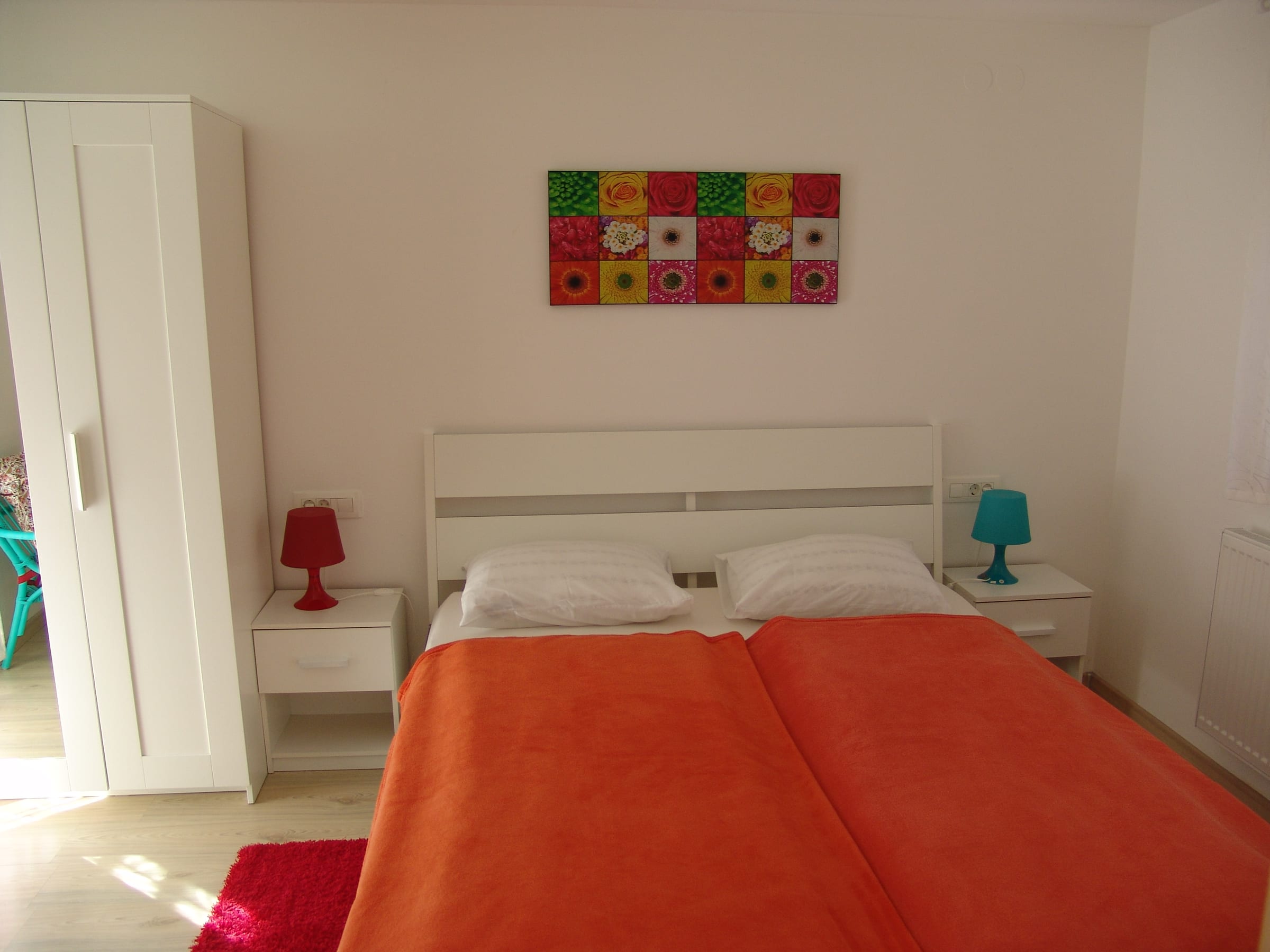 The double bed seen from the side, with a tall white wardrobe, a turquoise bedside lamp and colourful wall art.