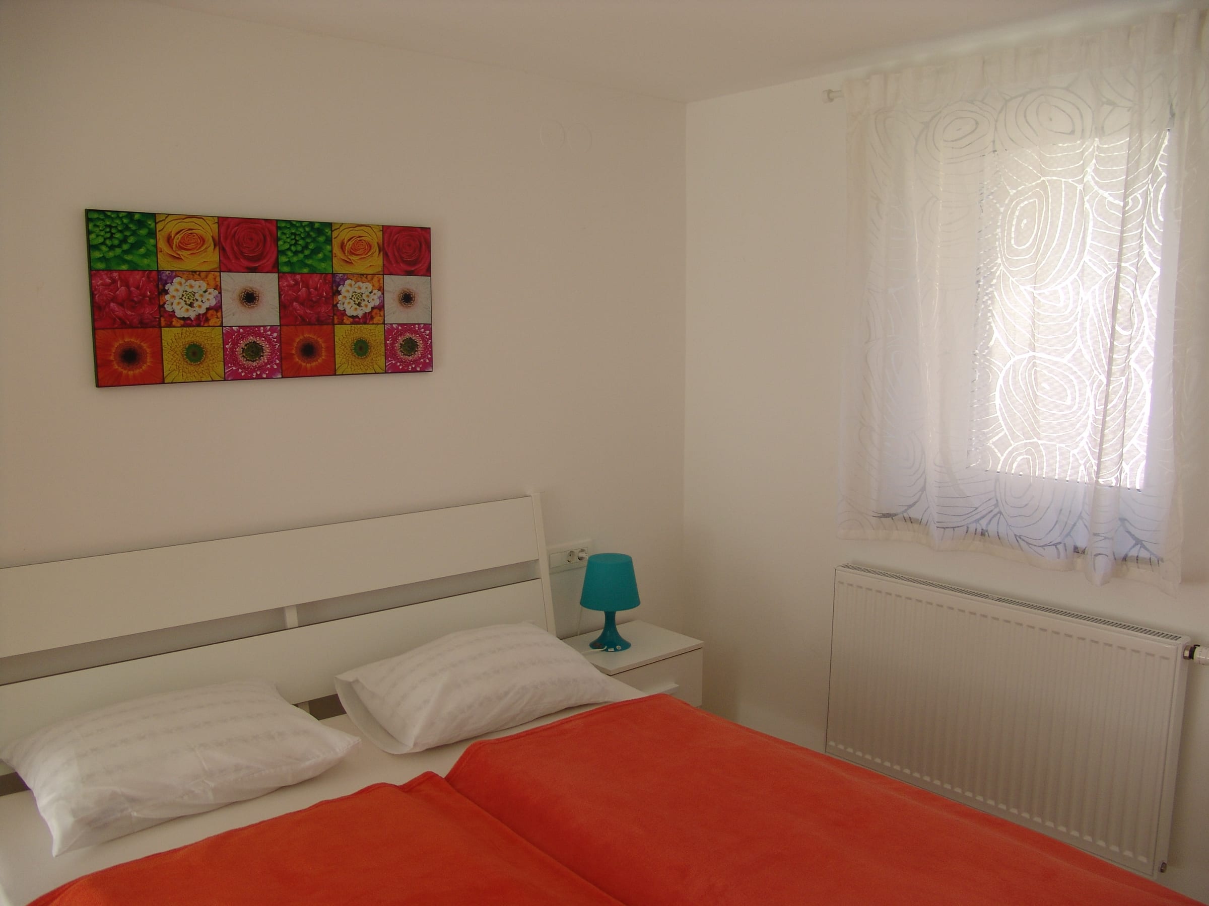 Double bed made up with a red cover, beside a curtained window and a framed panel of colourful printed squares.