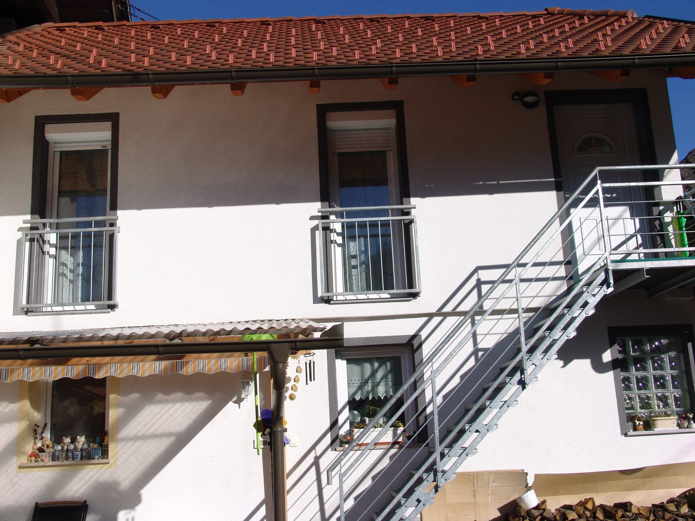 The house exterior in sunshine, with wooden shutters, balcony railings and an external metal staircase under a tiled roof.