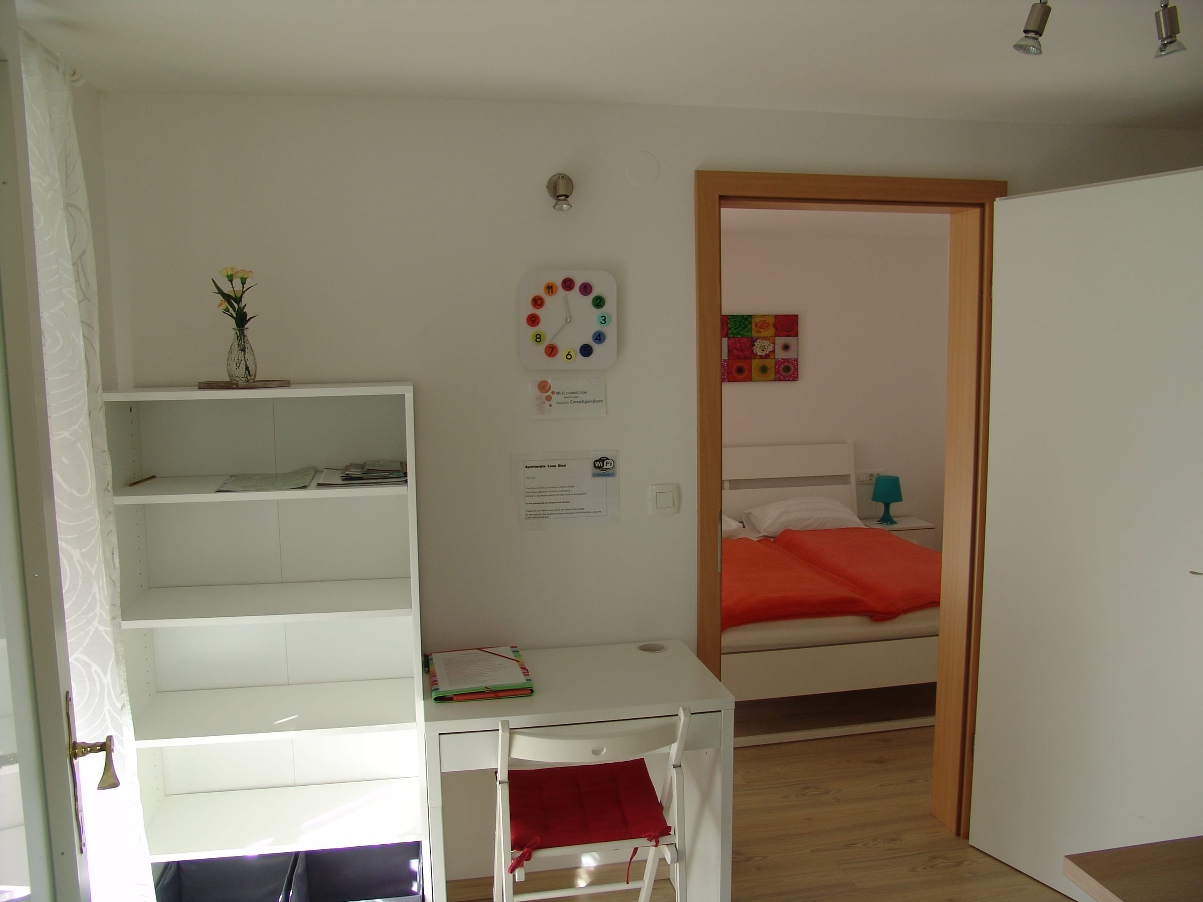 Looking from the entrance area, past open white shelving and a desk, through a doorway to the bed.