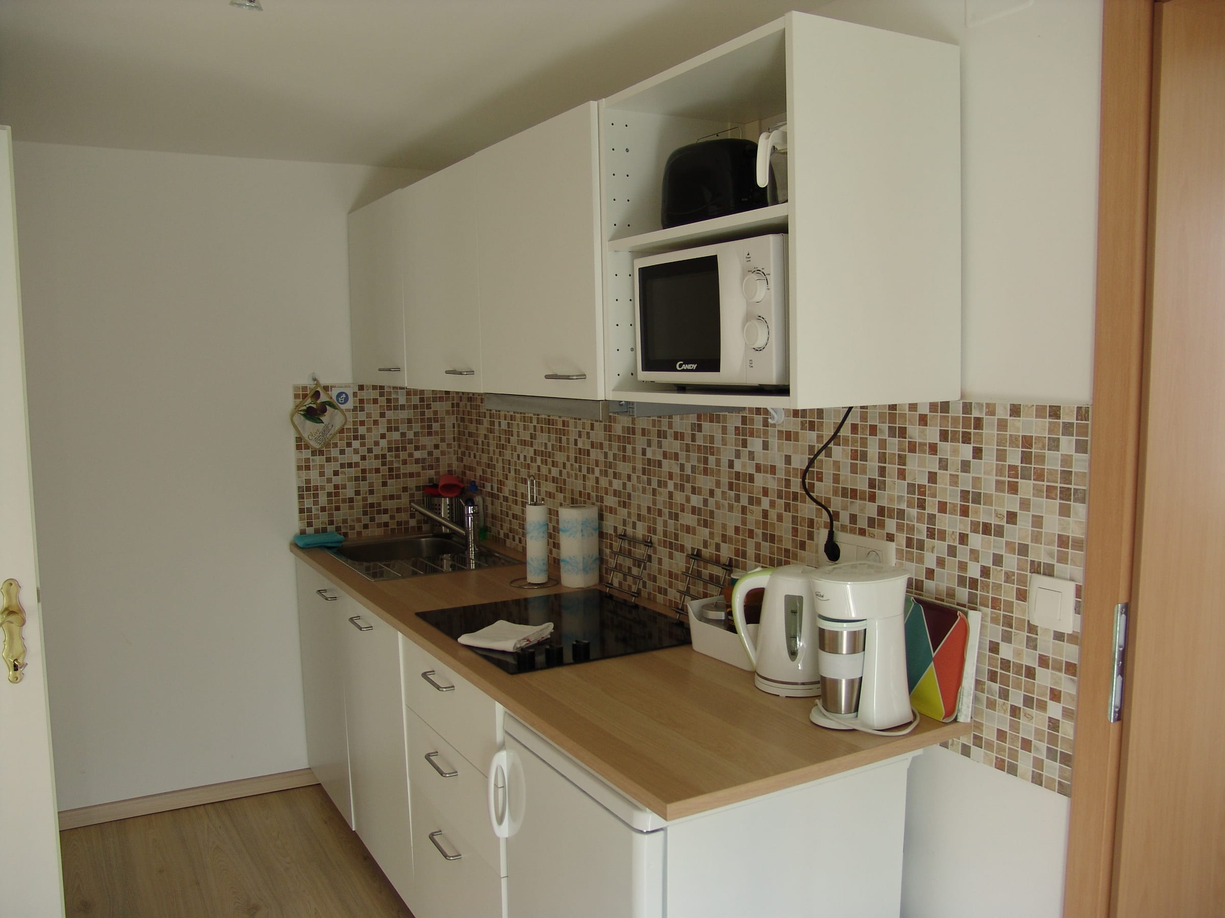 Kitchen corner with white units, a wooden worktop, mosaic tiled splashback, hob, microwave and electric kettle.