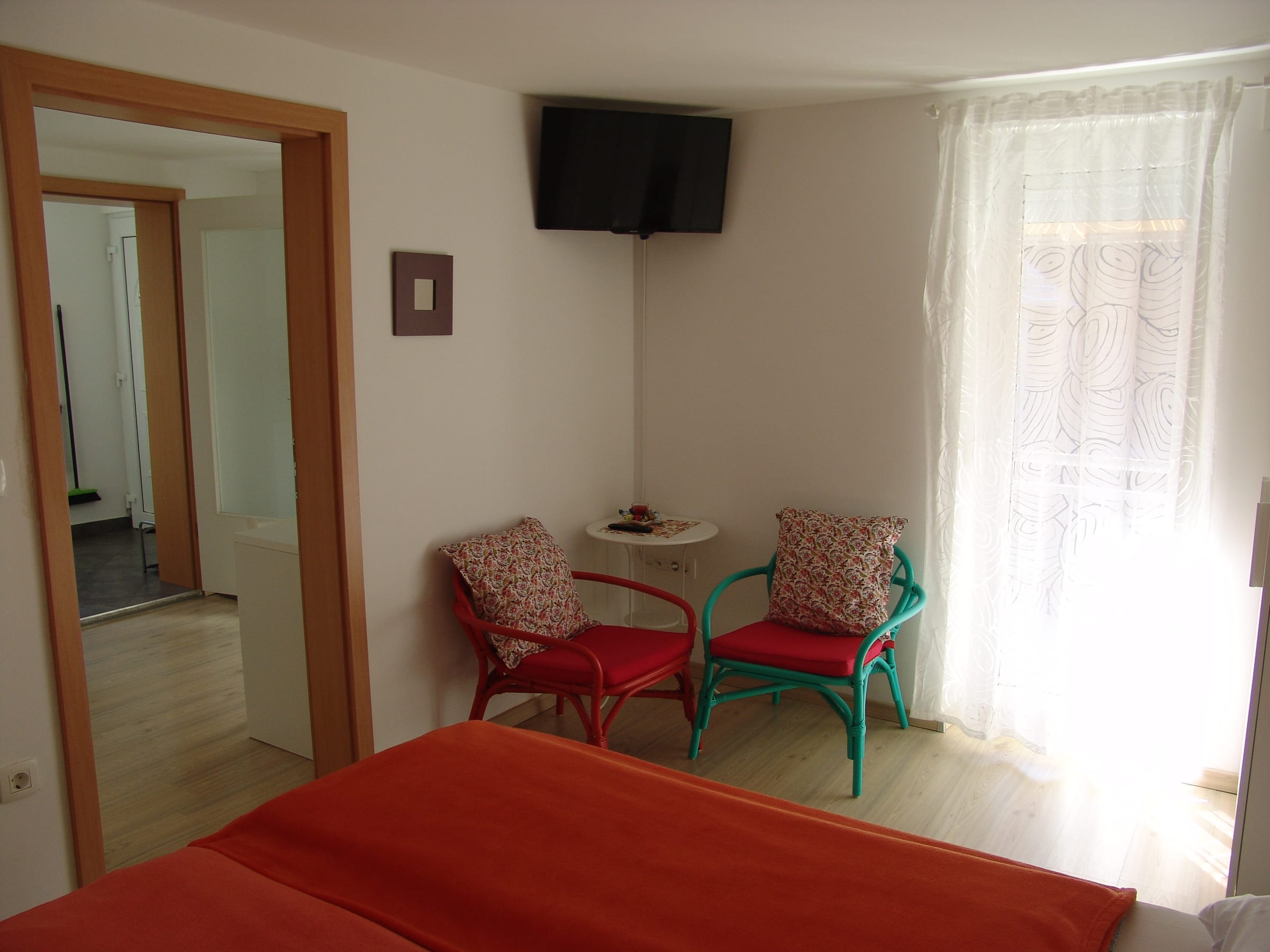 View across the foot of the bed towards the table, armchairs and window, with a doorway to the kitchen on the left.