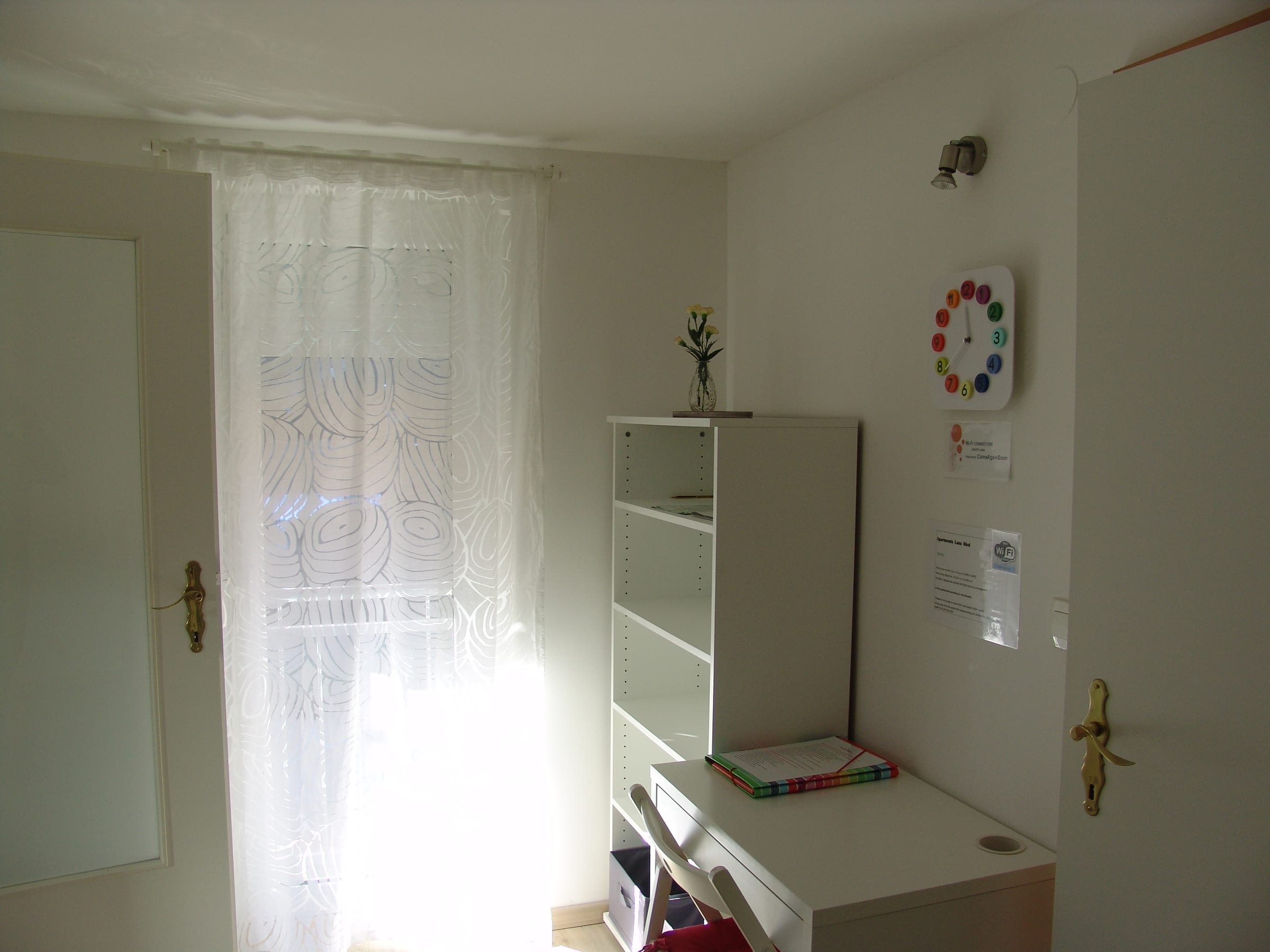A bright window with a white lace curtain beside open shelving and a wall clock.