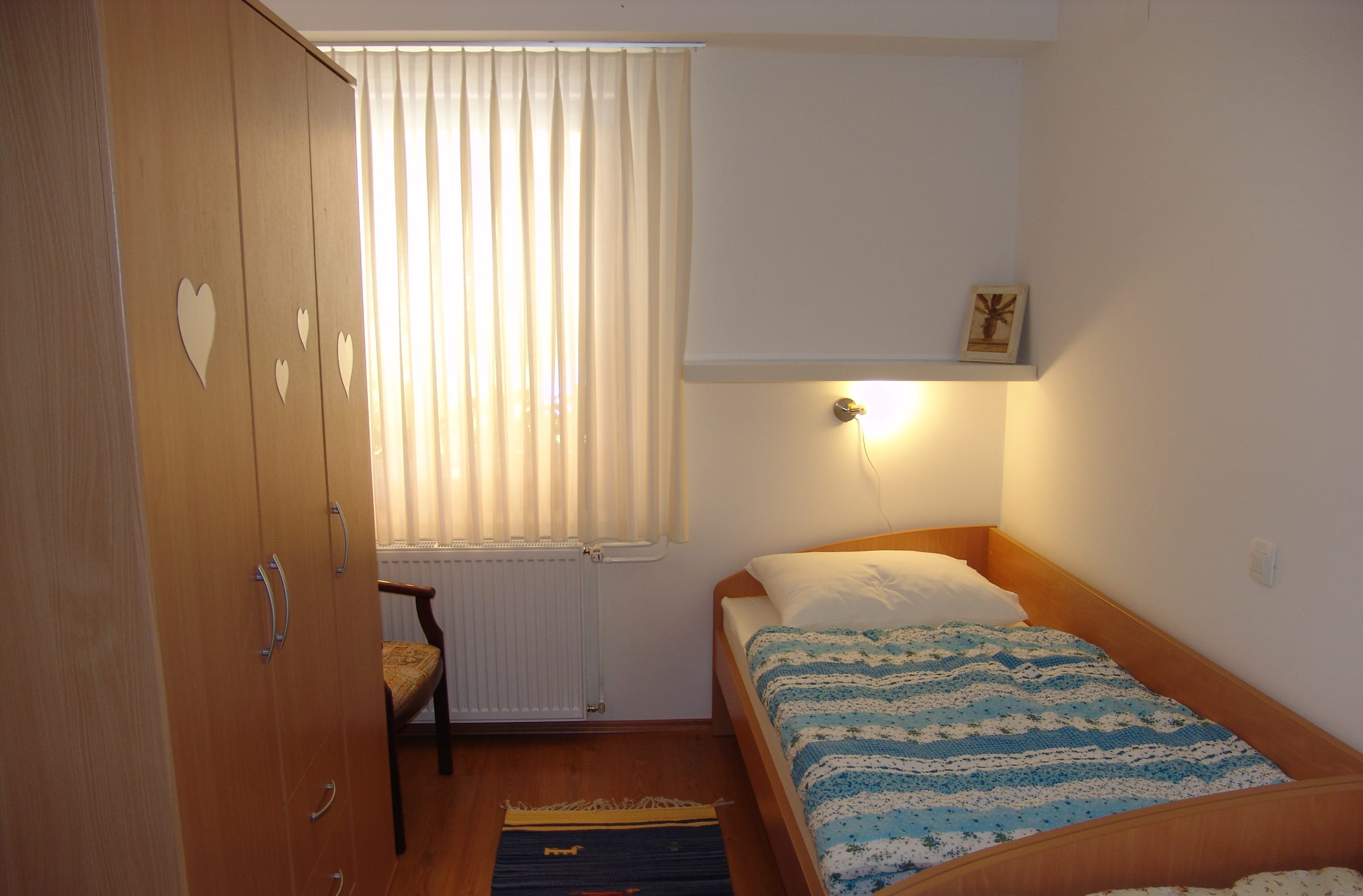 A single bed against a pale wall beside a tall wardrobe and a wall light.