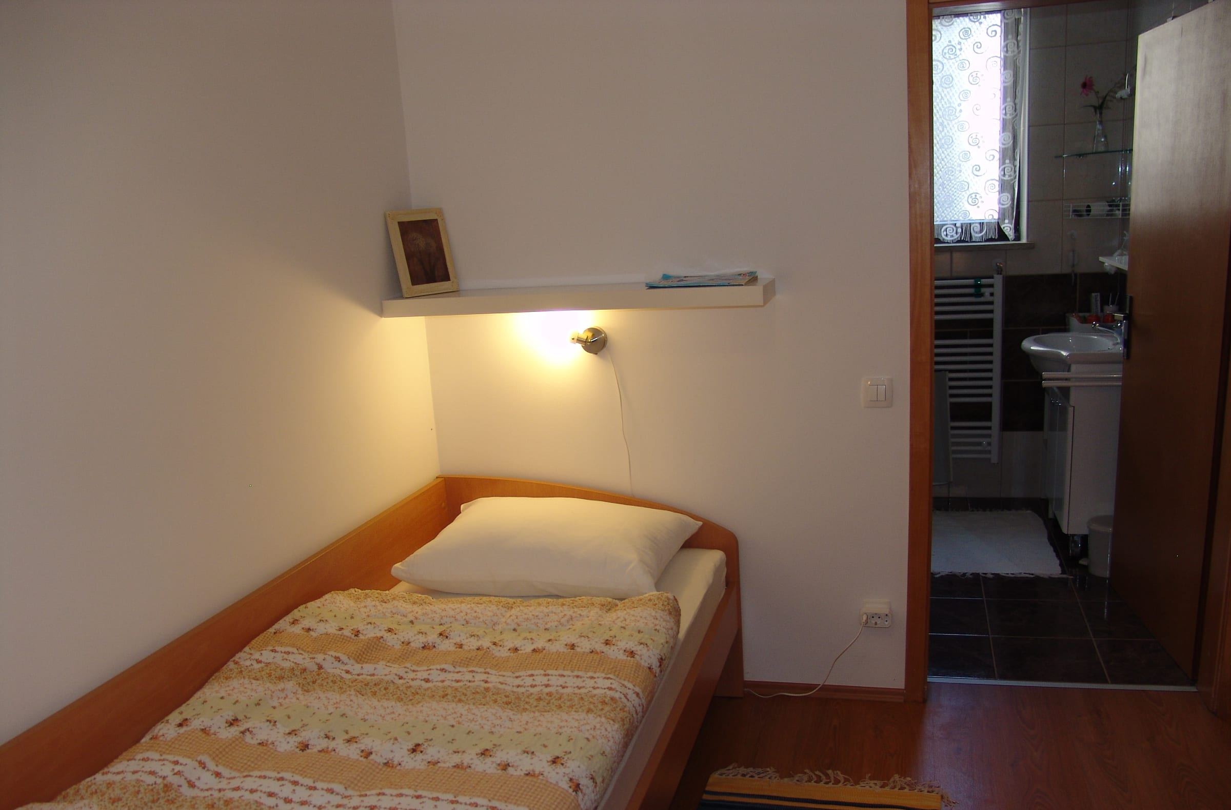 A single bed with a striped cover, a wall light above and a doorway through to the kitchen.
