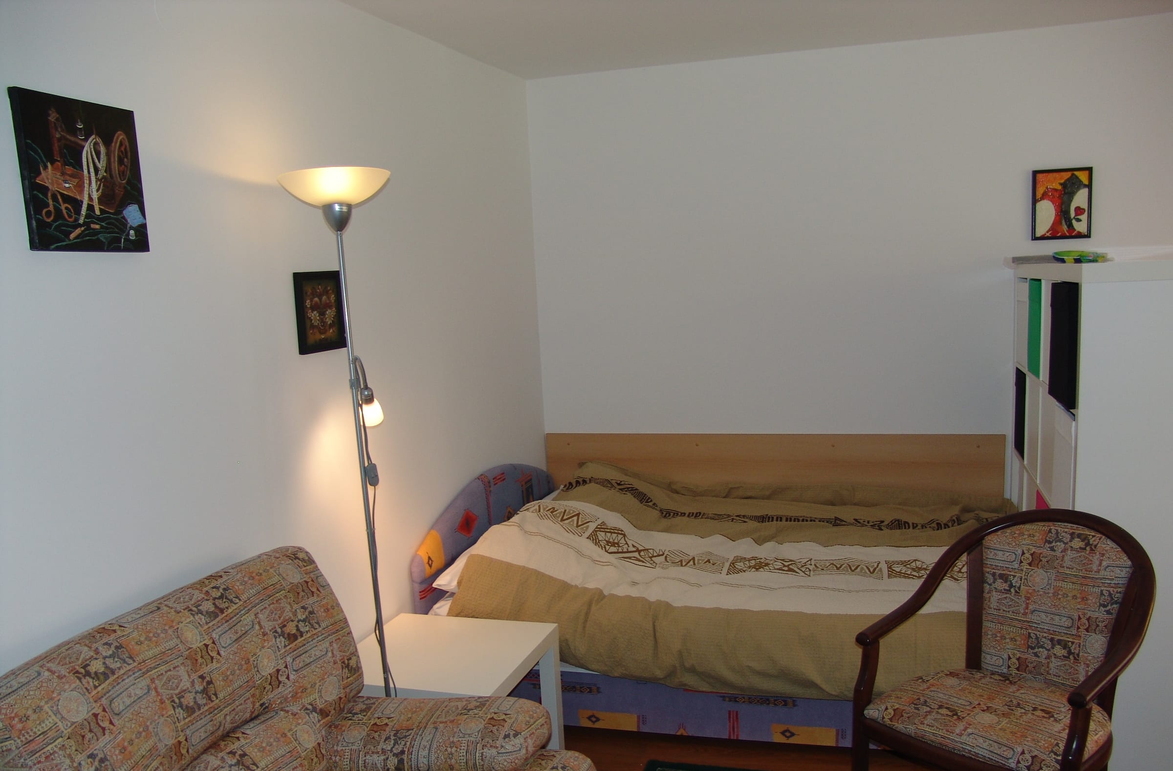 A living area with a sofa, an armchair, a floor lamp and a made-up bed along the far wall.