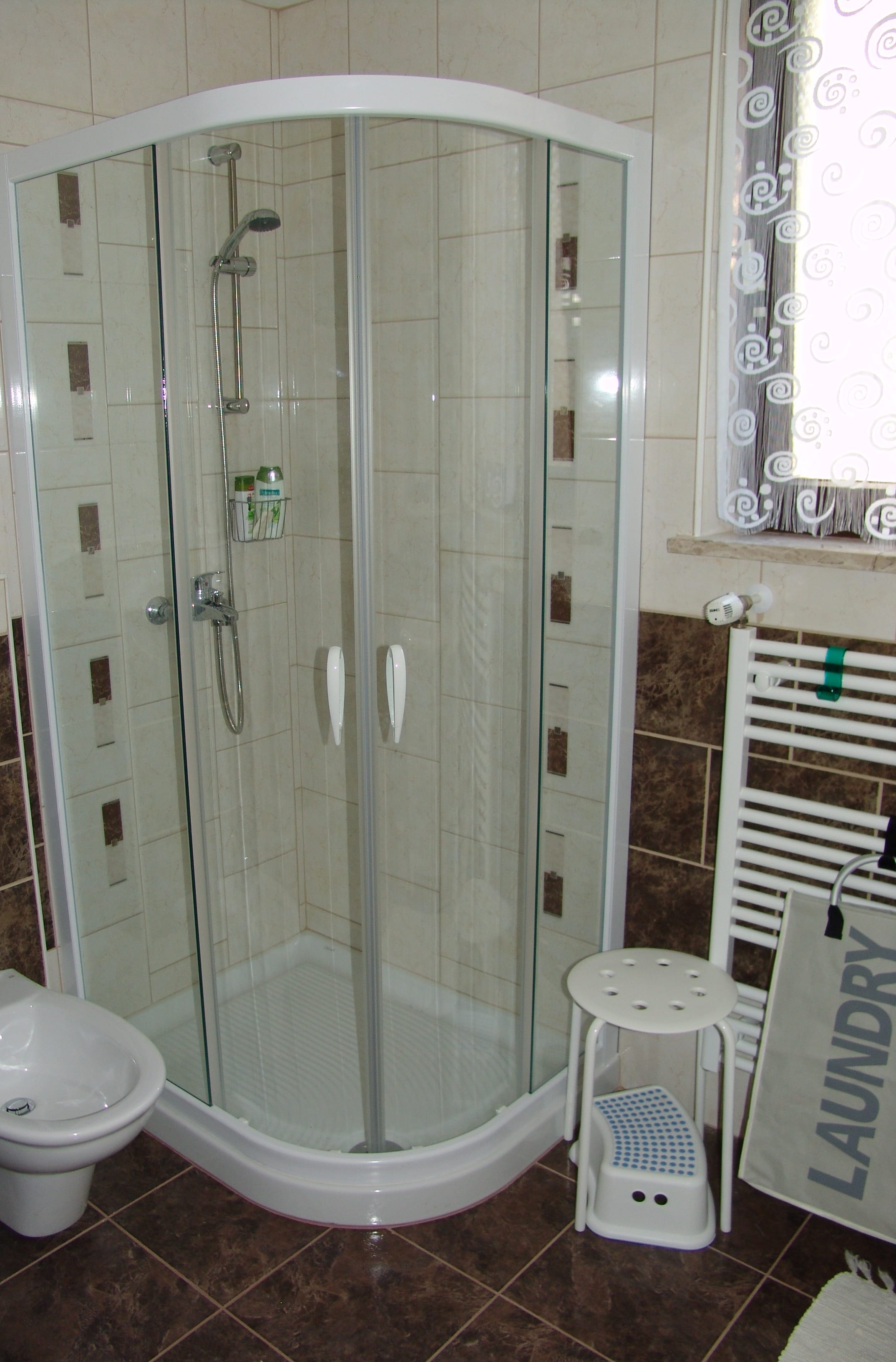 A quadrant shower cabin with a glass door, beside a heated towel rail and a curtained window.