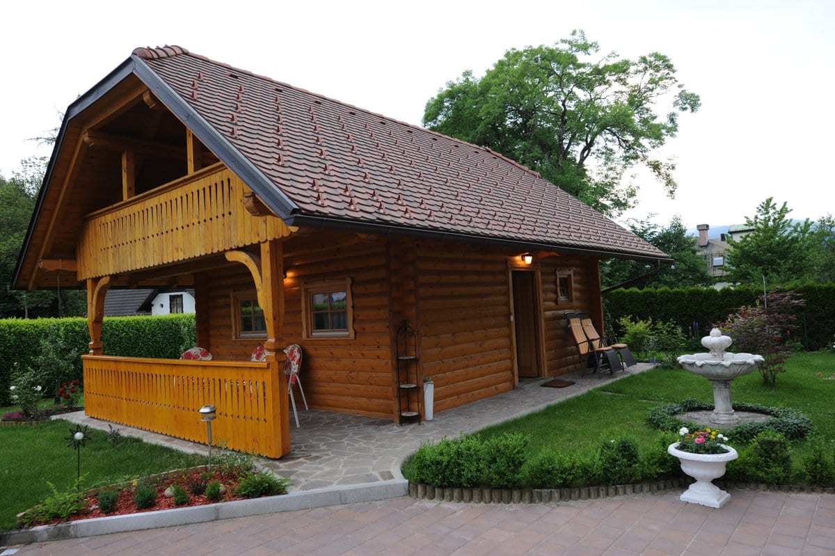 The timber chalet behind a small stone fountain, framed by clipped hedges and garden planting.