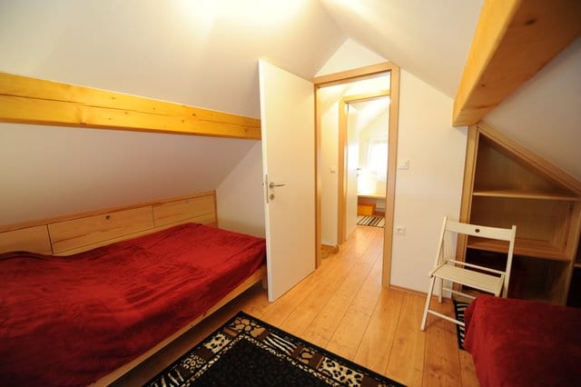 A bedroom with red covers and a pine wall, looking through an open door to the landing.