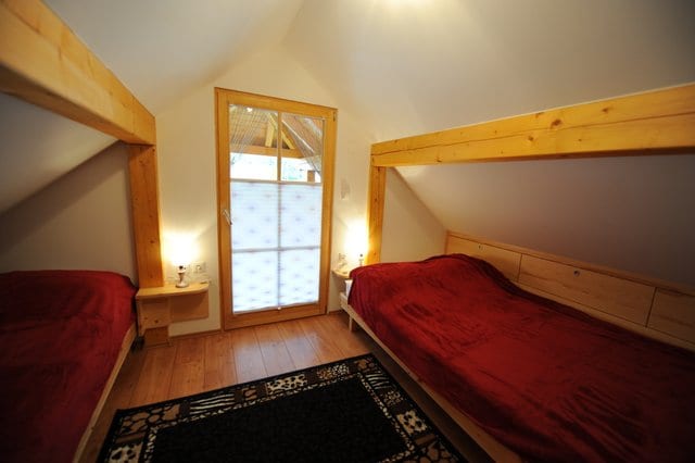 A bedroom with dark red bed covers beneath a pine ceiling and a window with timber surrounds.
