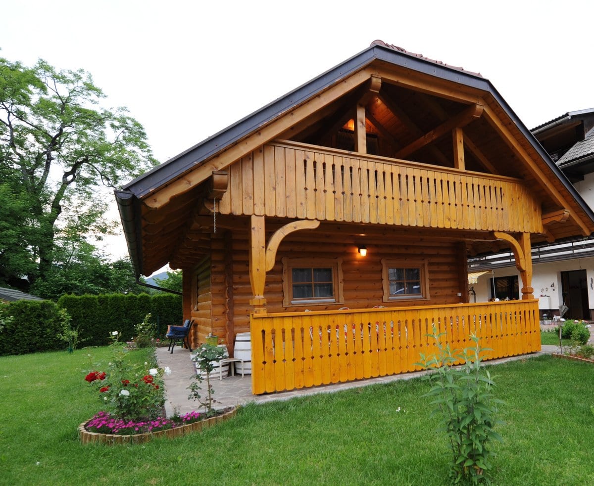 The timber chalet with its covered veranda and carved balcony, seen across a mown lawn and flower beds.