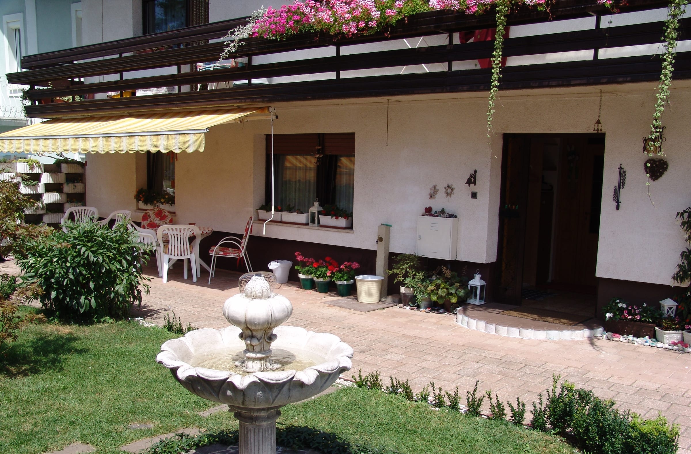 A stone fountain on the lawn in front of the house, with awnings shading the ground-floor terrace.