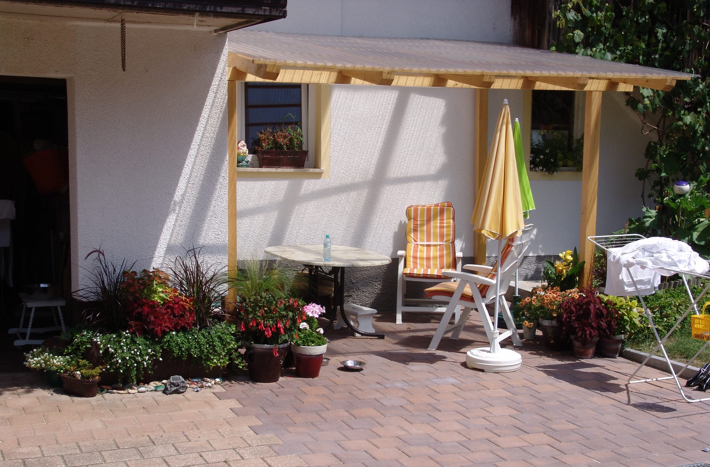 A timber pergola beside the house with a parasol, garden chairs and flower pots on the paving.
