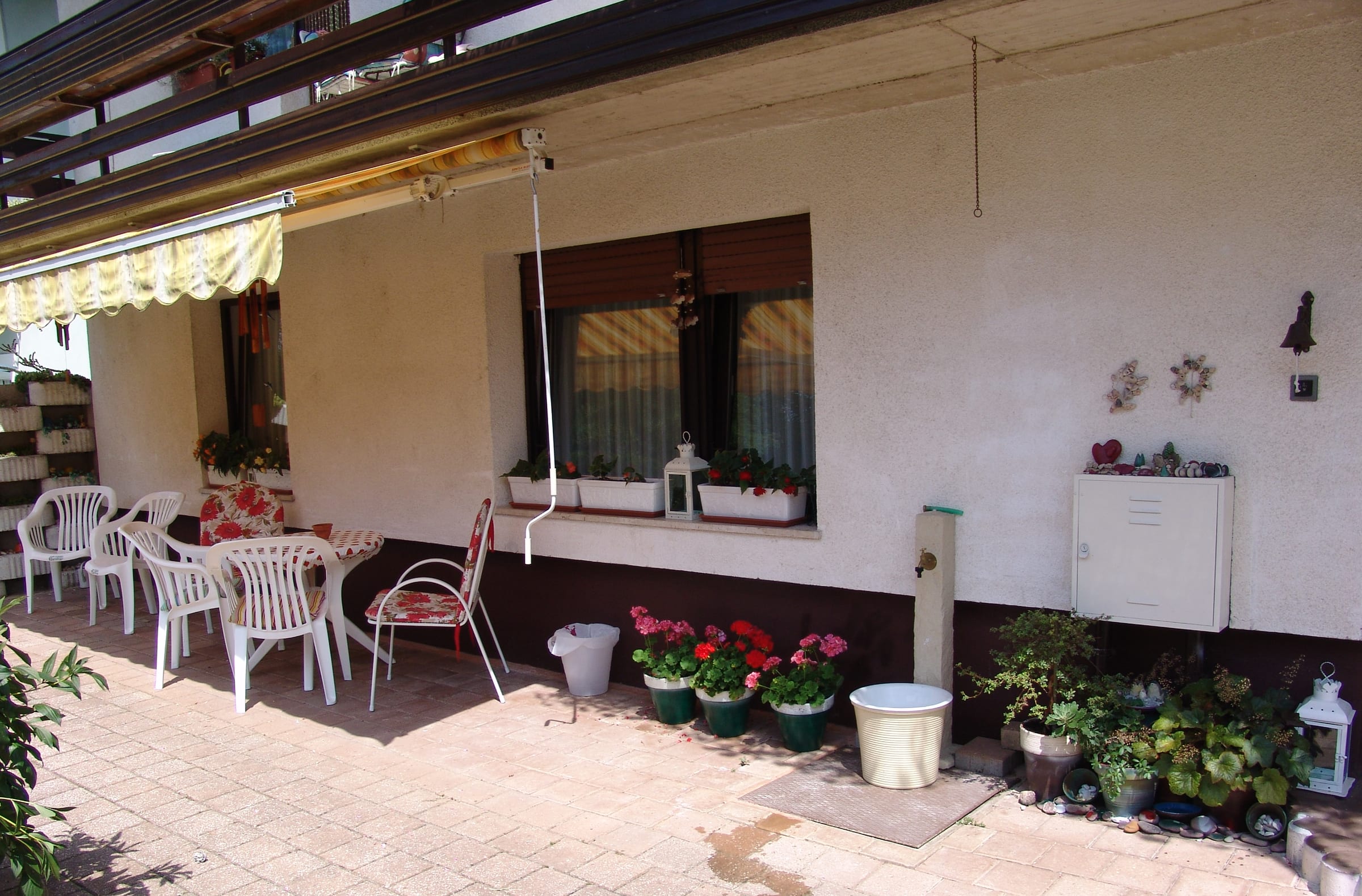 A shaded ground-floor terrace with garden chairs and potted flowers along the wall of the house.